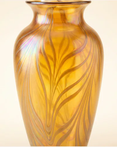 An Art Nouveau iridescent glass vase with trailing organic decoration, lit from behind on a warm paper plate.