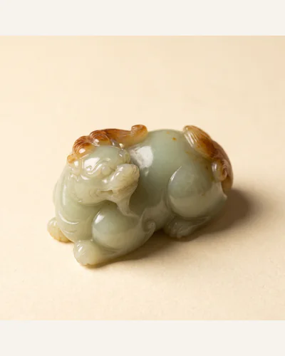 A pale celadon jade carving of a recumbent beast with russet inclusions, photographed under macro lighting.