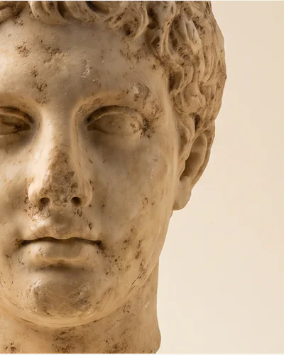 A weathered marble head of a youth on a steel display mount, photographed on a warm paper plate. — burial patina.