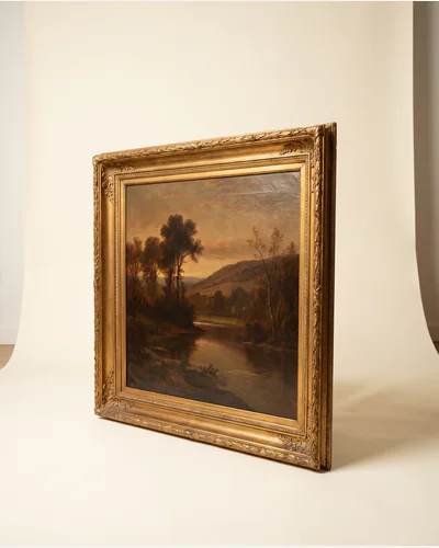 An antique oil landscape of a river valley at dusk in a carved gilt frame, photographed upright on a warm paper plate.