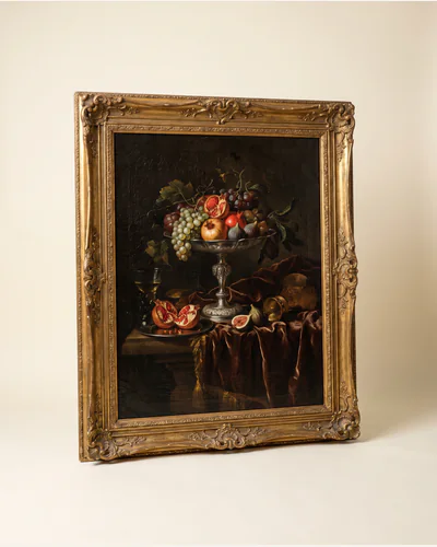 A dark Flemish still life of fruit, a silver tazza and drapery in a moulded gilt frame, photographed on a warm paper plate.